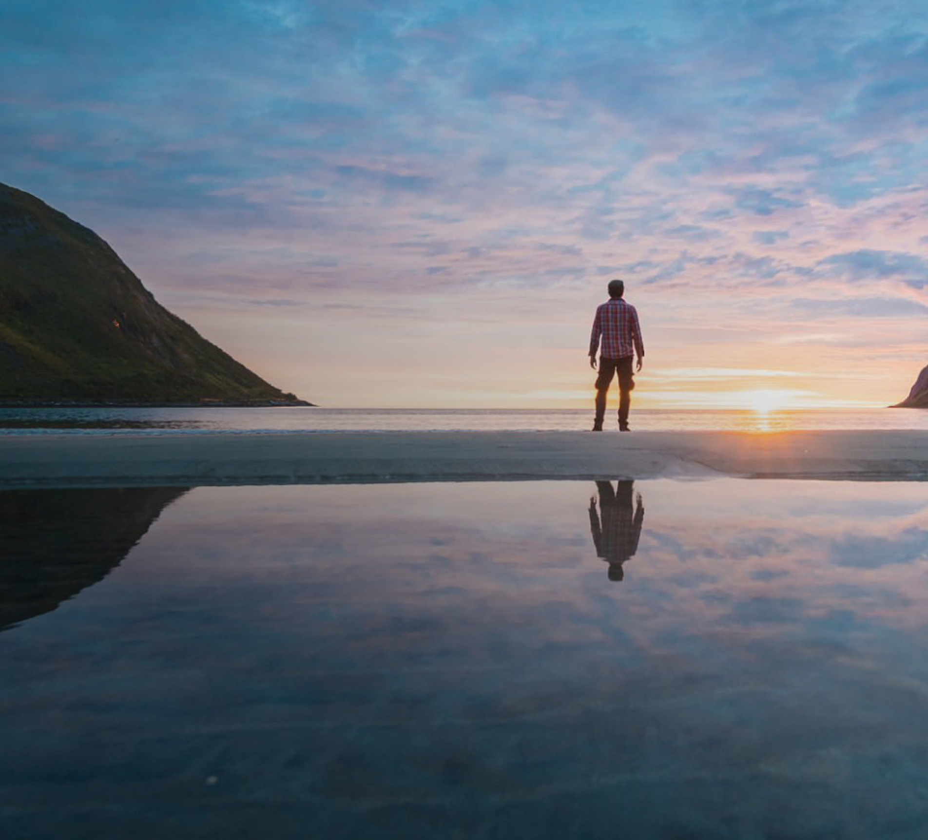 Hombre de pie en una playa al atardecer, contemplando el horizonte con el reflejo en el agua
